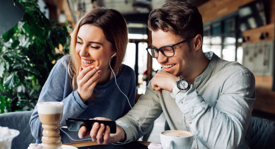 A man and a woman laugh while listening to something on a phone and drinking coffee